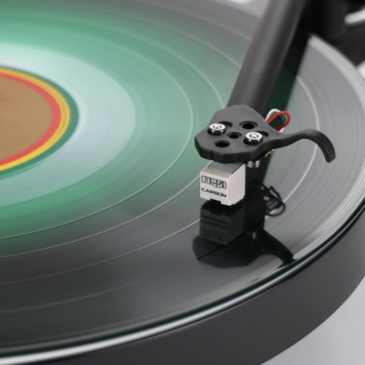 Close-up of a black turntable needle cartridge, resting on a vinyl record with a green, yellow, and red label visible in the background.