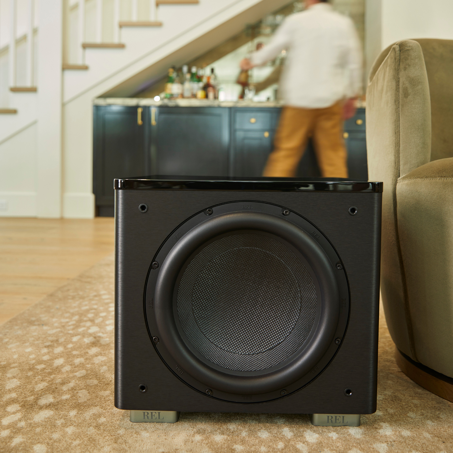 A black REL audio subwoofer placed on a patterned beige rug in a living room setting, with a blurred background showing a staircase, a person at a bar, and a sofa.