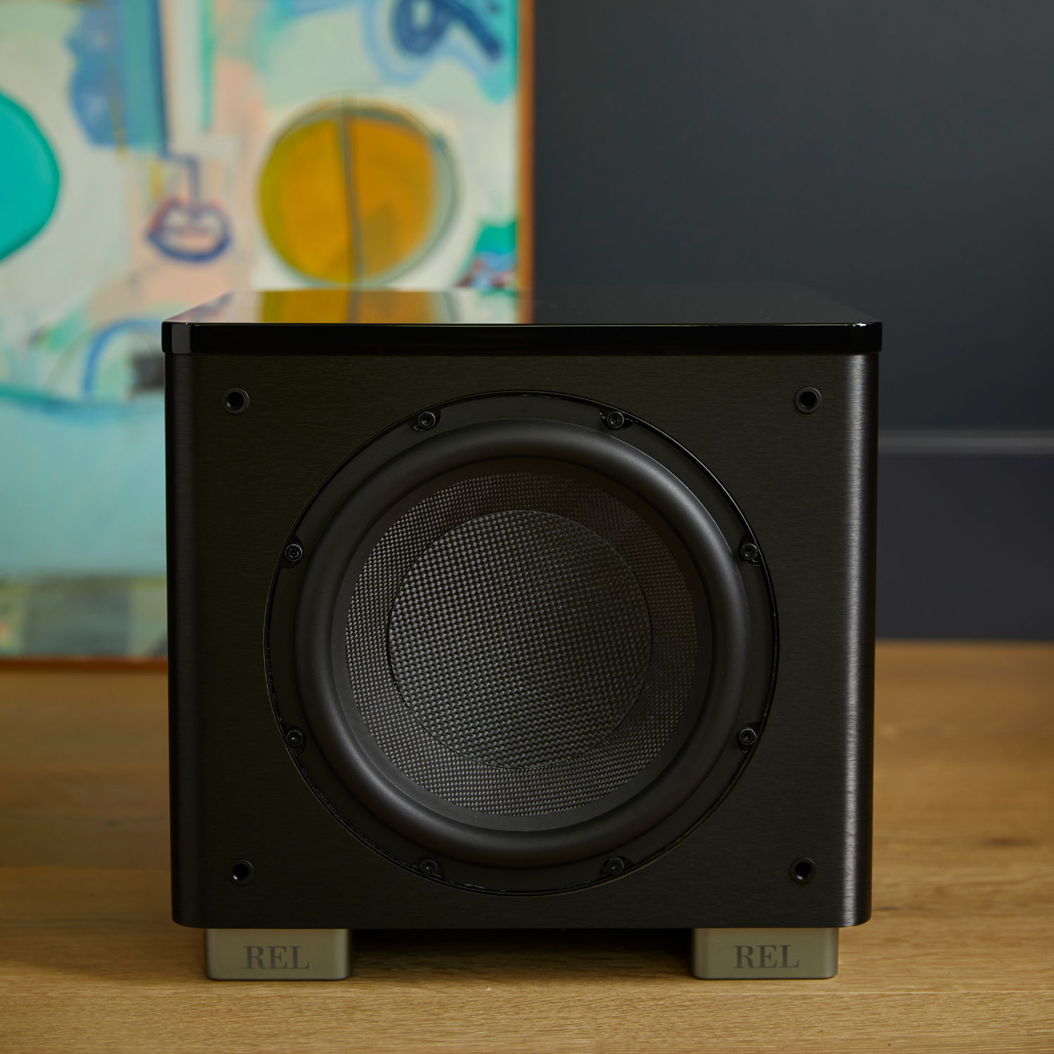 A black REL subwoofer with a circular driver and gray feet, sitting on a wooden surface with a dark gray wall and colorful abstract art in the background.