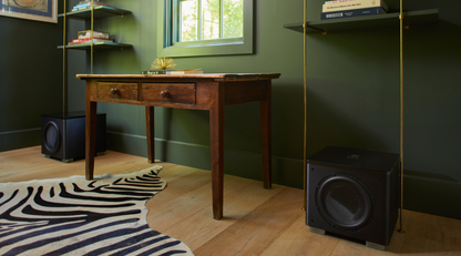 A dark wood writing desk in a home office setting with olive green walls, a zebra-print rug, and black subwoofers on the floor.