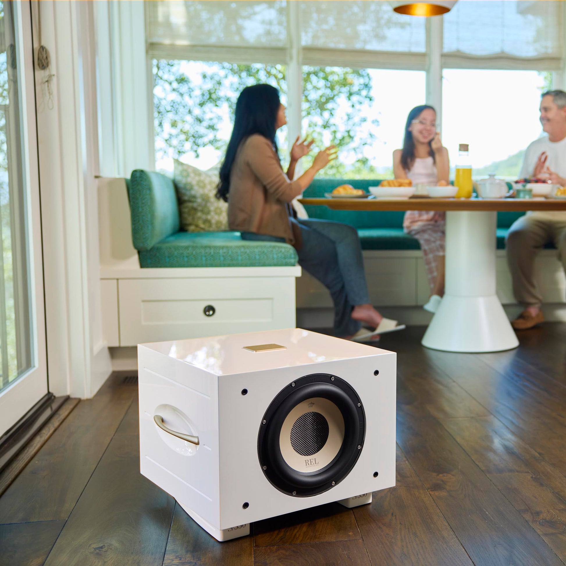 White subwoofer on a wooden floor with people sitting at a table in the background