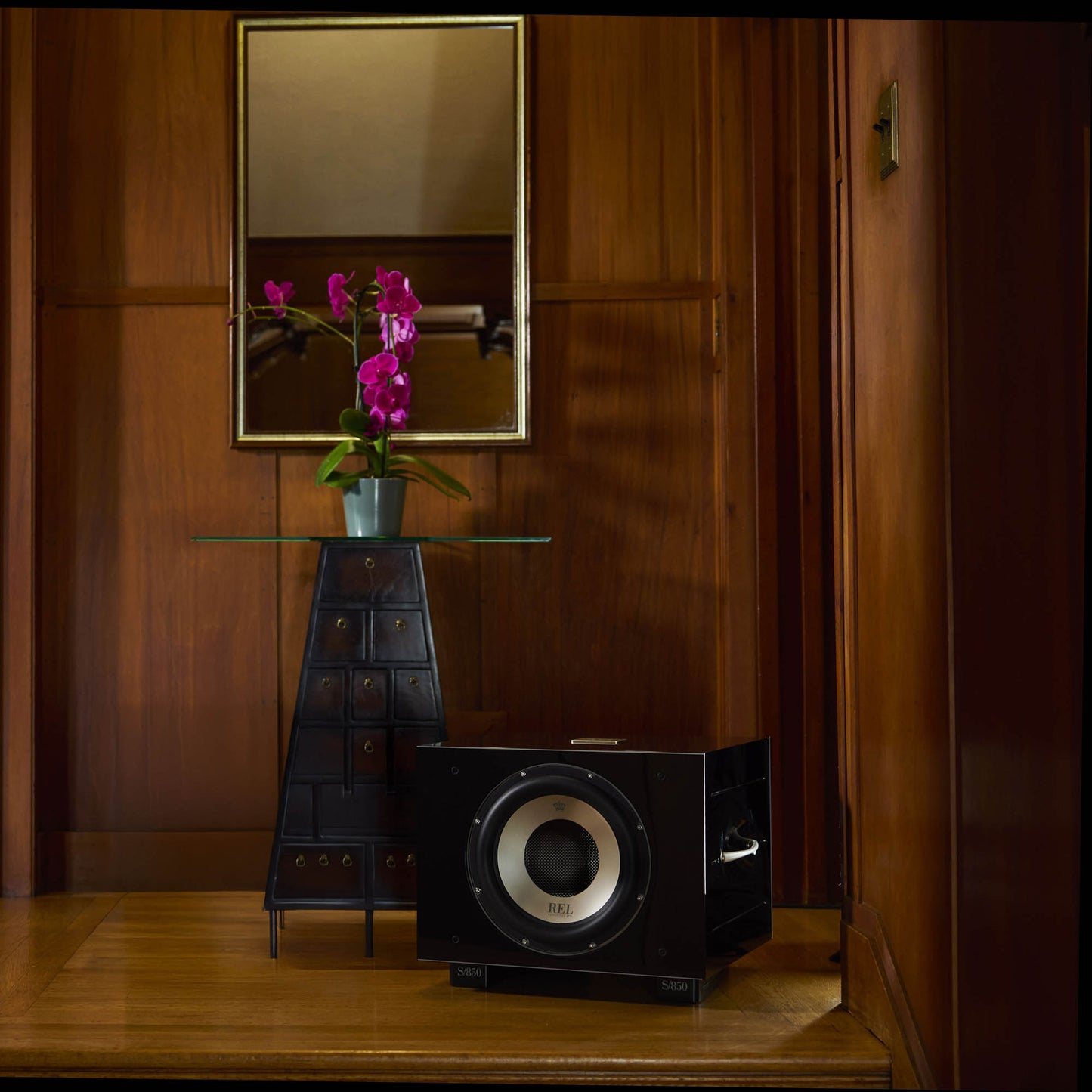 Black subwoofer on a wooden floor with a mirror reflecting flowers in a room.