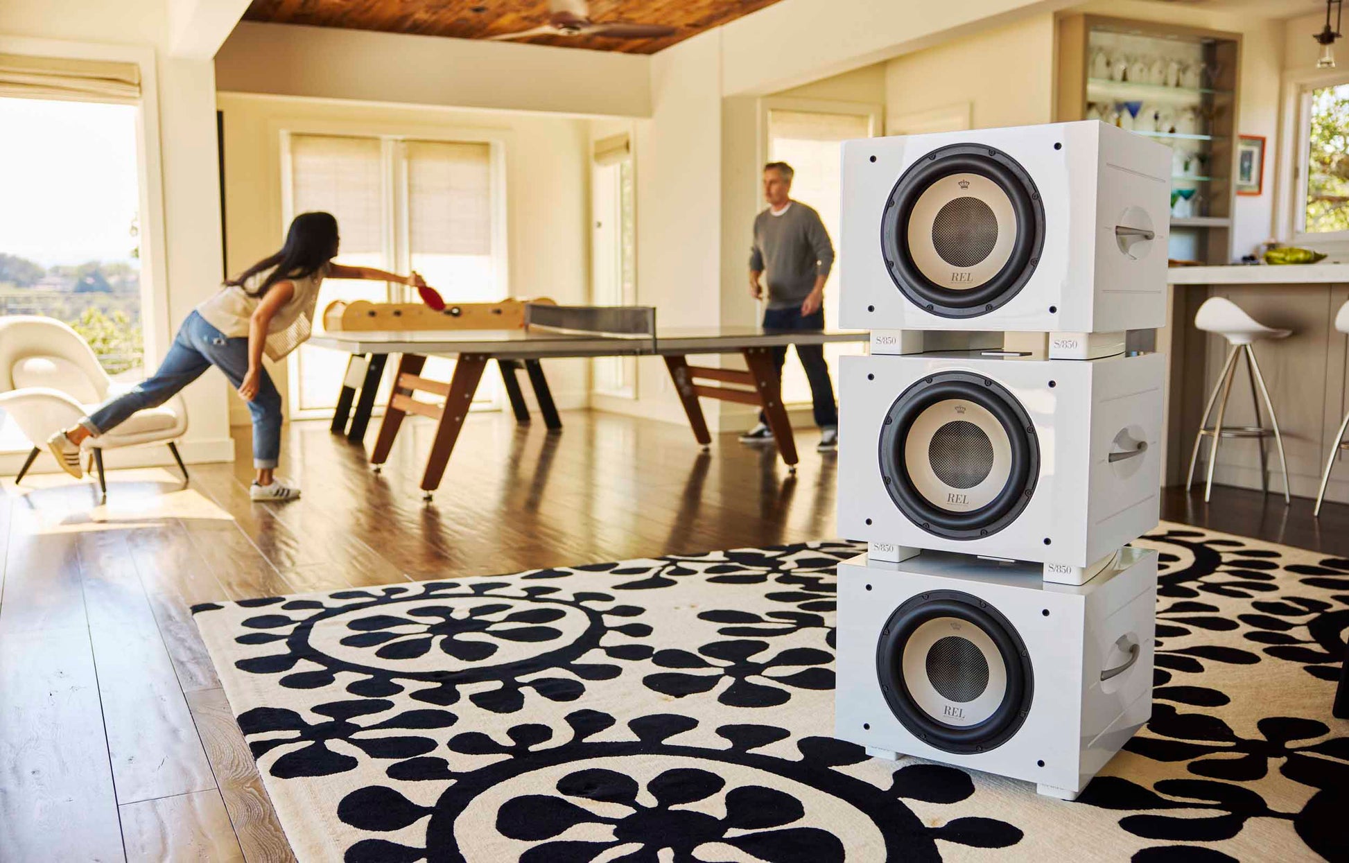 Stack of white REL S/850 Subwoofer on a patterned rug with people in the background