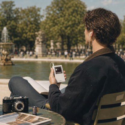 A person sitting outdoors by a lake in a park, holding a white portable music player with wired earphones. The scene includes a table with a camera and magazine in the foreground and trees and a fountain in the background.