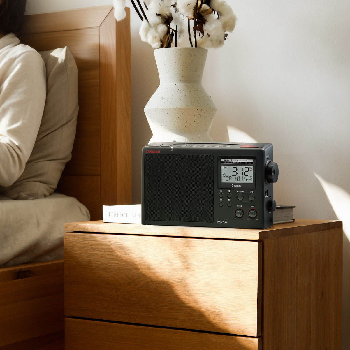 Digital clock radio on a wooden nightstand next to a bed with a vase of flowers.