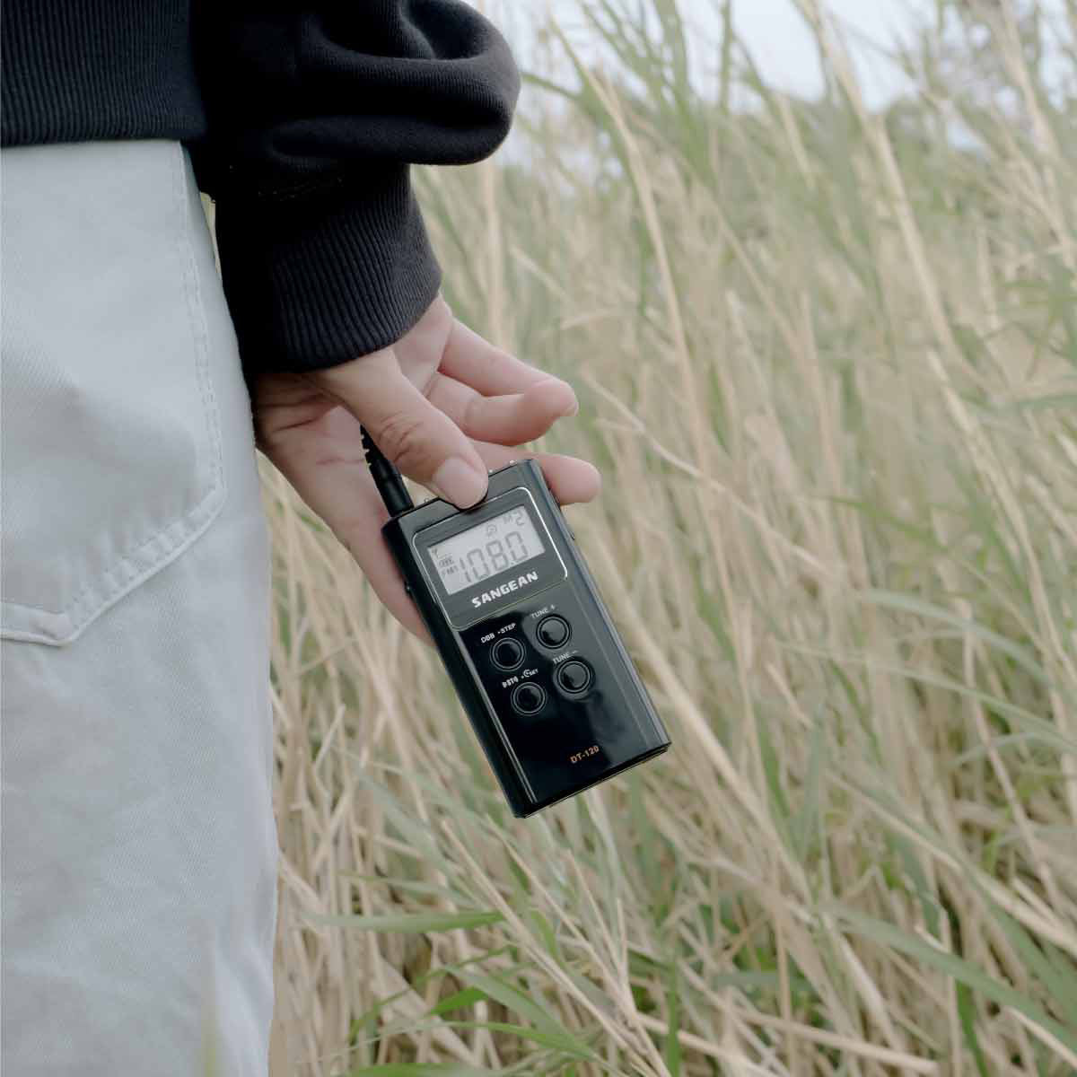Person holding a Sangean DT-120 AM/FM Stereo Pocket Radio in a grassy outdoor setting