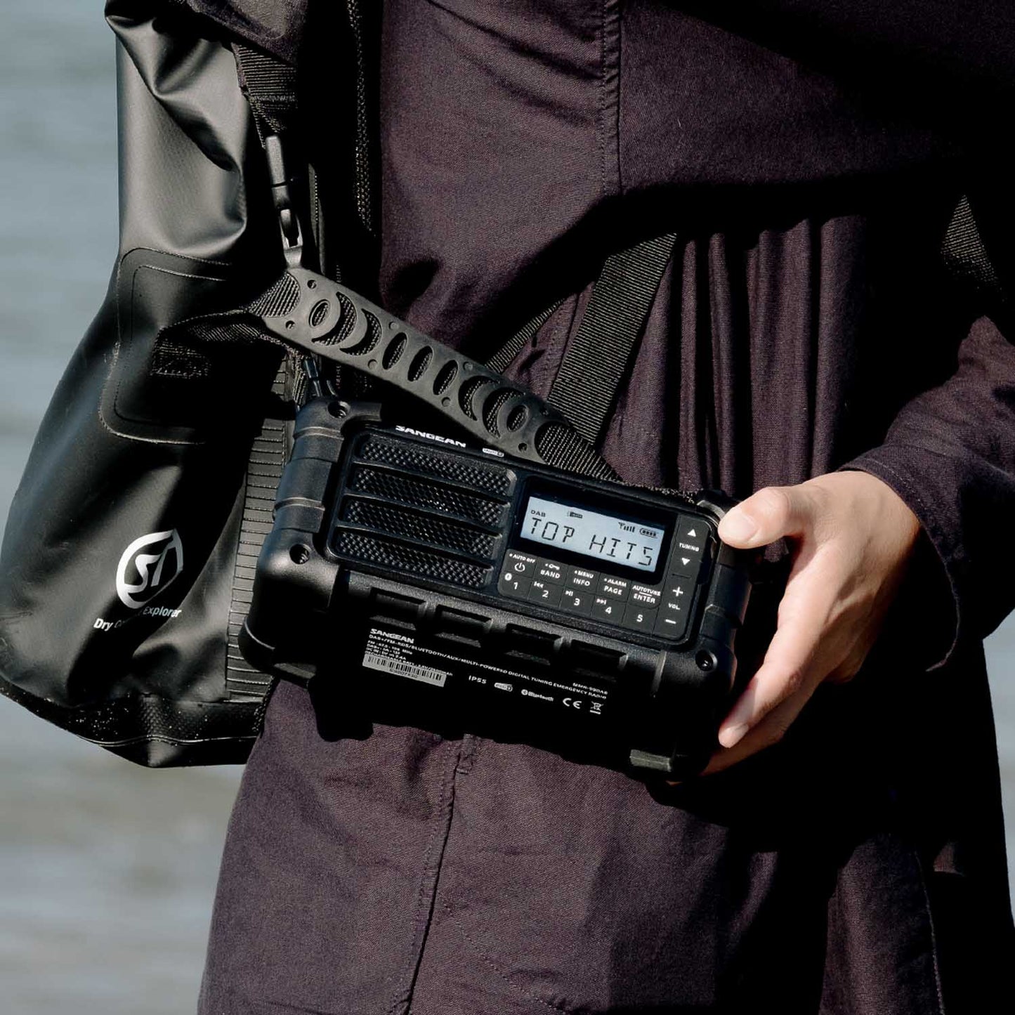 Person holding a black radio device with a digital display, wearing a dark jacket and carrying a black bag.