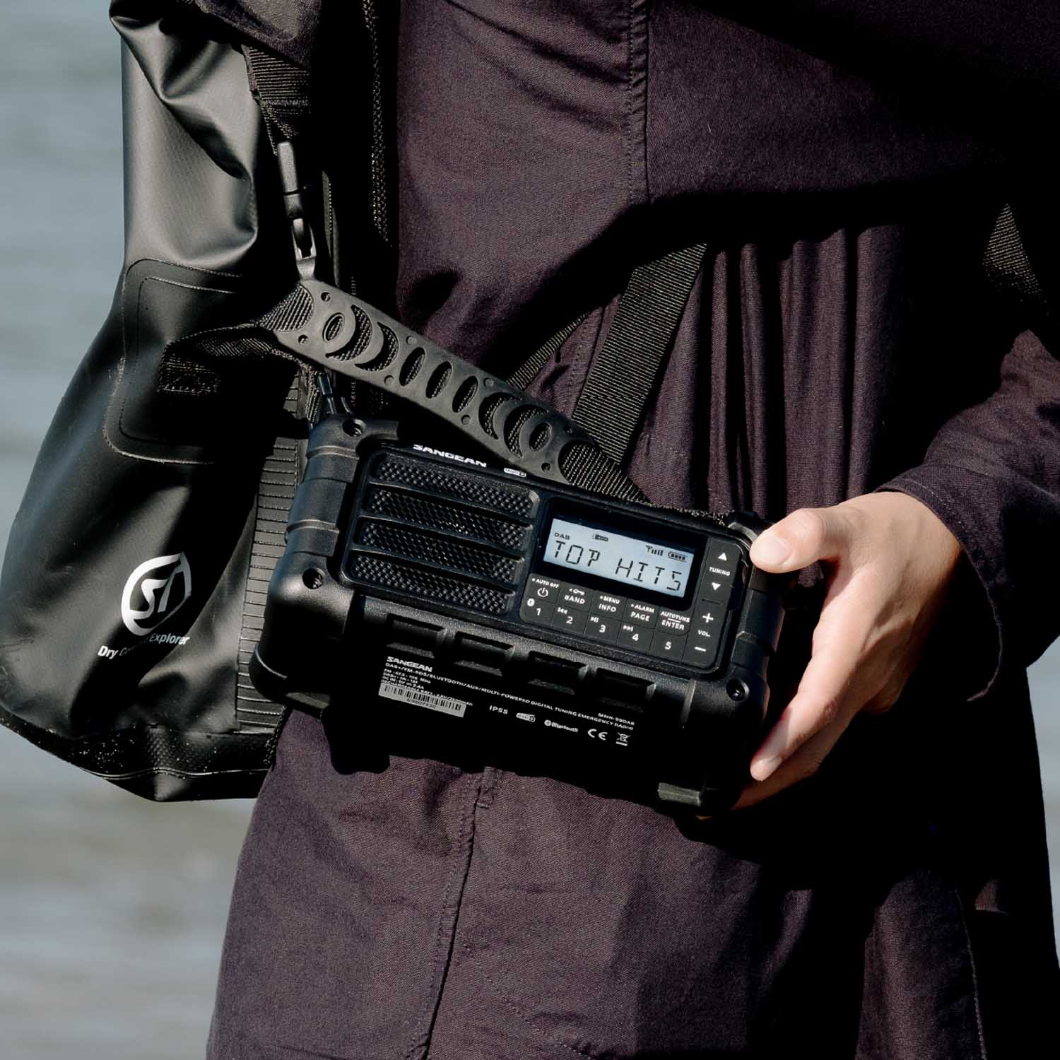 Person holding a black radio device with a digital display, wearing a dark jacket and carrying a black bag.