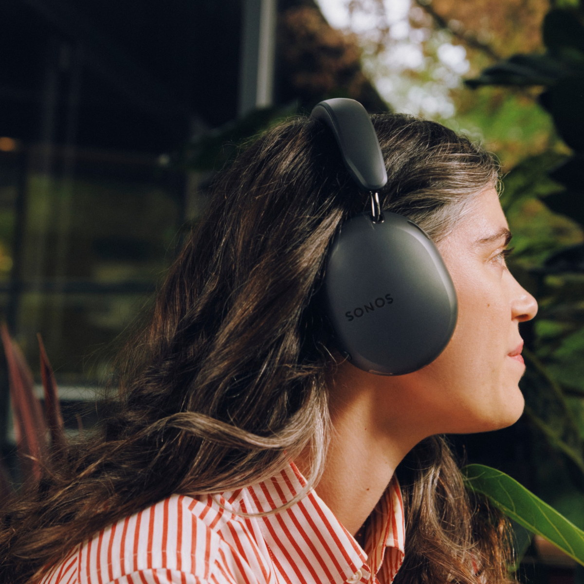 A woman in profile wearing black over-ear headphones with the brand name 'SONOS' visible on the side, set against a blurred background of green foliage and glass.