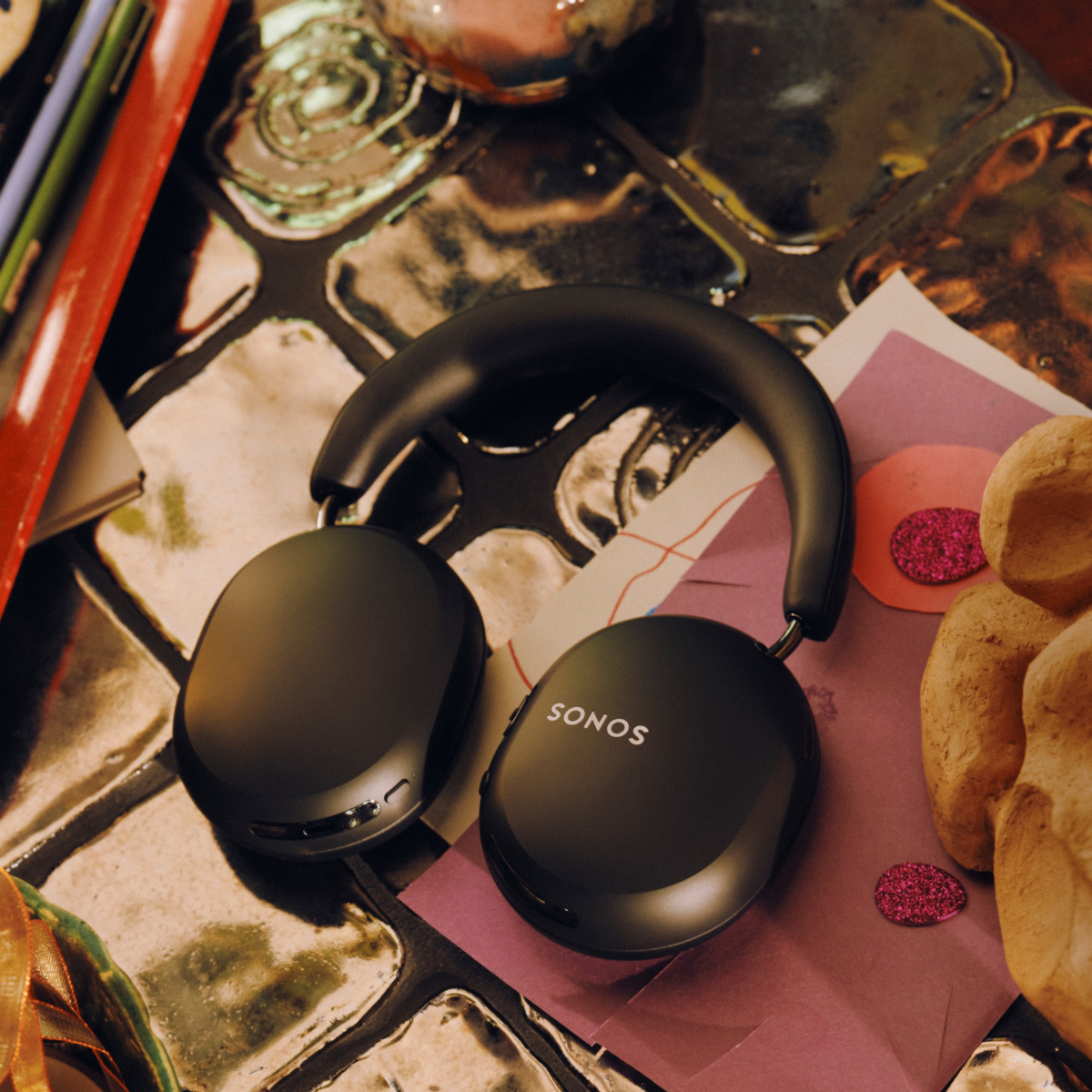Black over-ear wireless headphones with the SONOS logo on the earcups, resting on a pink and white patterned sheet of paper on a reflective tiled surface.