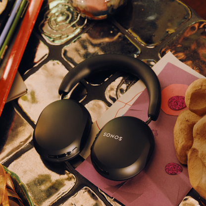 Black over-ear wireless headphones with the SONOS logo on the earcups, resting on a pink and white patterned sheet of paper on a reflective tiled surface.