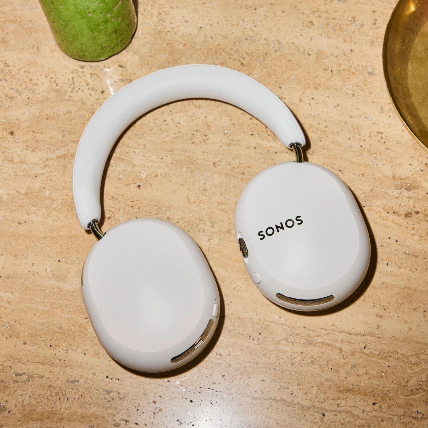 White over-ear wireless headphones with the SONOS logo printed on the right earcup, resting on a beige stone surface.