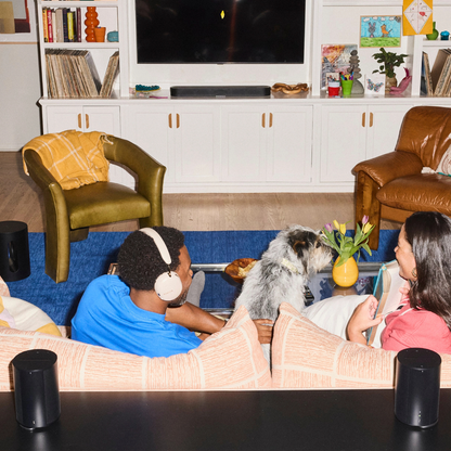 A cozy living room scene with two people sitting on a patterned sofa, a dog at the coffee table, and a TV and shelves in the background. The image appears to showcase smart home or audio devices, including black cylindrical speakers on the table in the foreground and white over-ear headphones worn by the person on the couch.