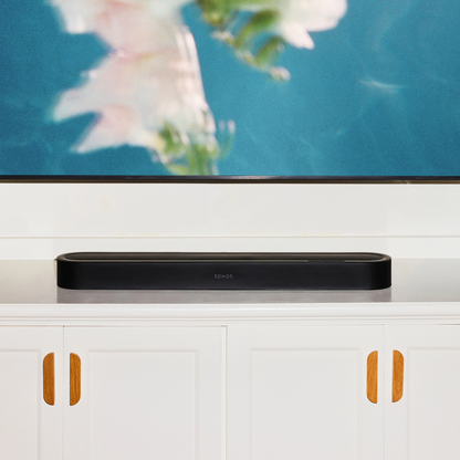 A sleek black Sonos soundbar sits on a white media console in front of a large, blurred TV displaying a blue floral scene.