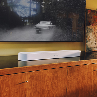 A white rectangular soundbar speaker sits on a glossy dark cabinet in a real interior setting, with a large TV screen above showing a car scene and a wooden cabinet below.