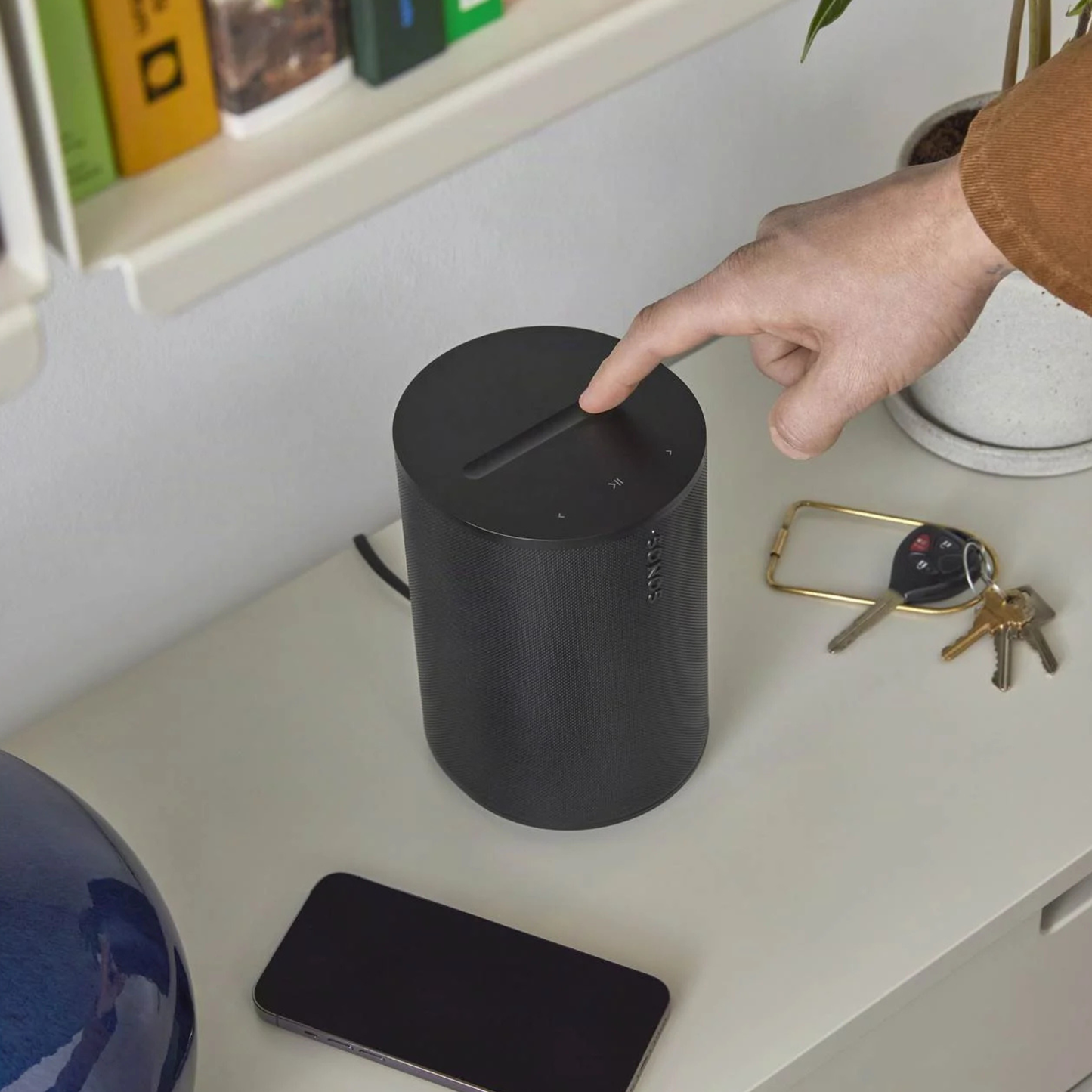 Person playing a black Sonos Era 100 on a desk with keys and phone nearby