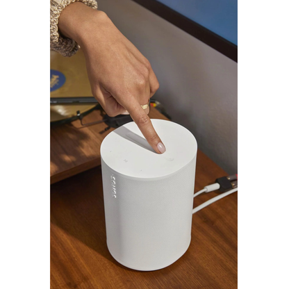 Hand pressing a white Sonos Era 100 on a wooden surface with a blurred background