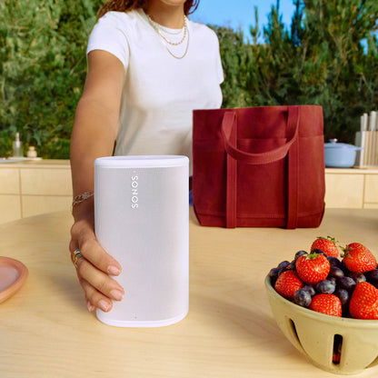 Person holding a white Sonos Play next to a red bag and bowl of fruit on a table outdoors.
