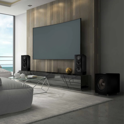 A modern living room setup featuring a large wall-mounted TV, a glossy black media console, two speakers, and a subwoofer on the floor, with a light gray sofa and ocean view in the background.