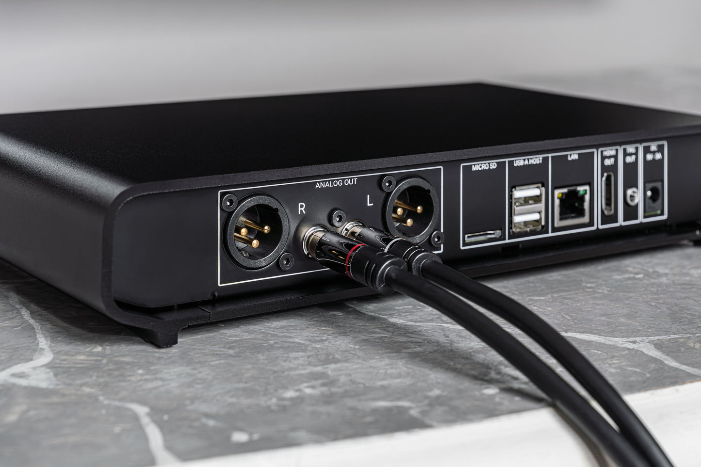 Close-up of the rear panel of a black audio device with XLR outputs, USB-A and USB-C ports, and connected audio cables, resting on a gray marble surface.