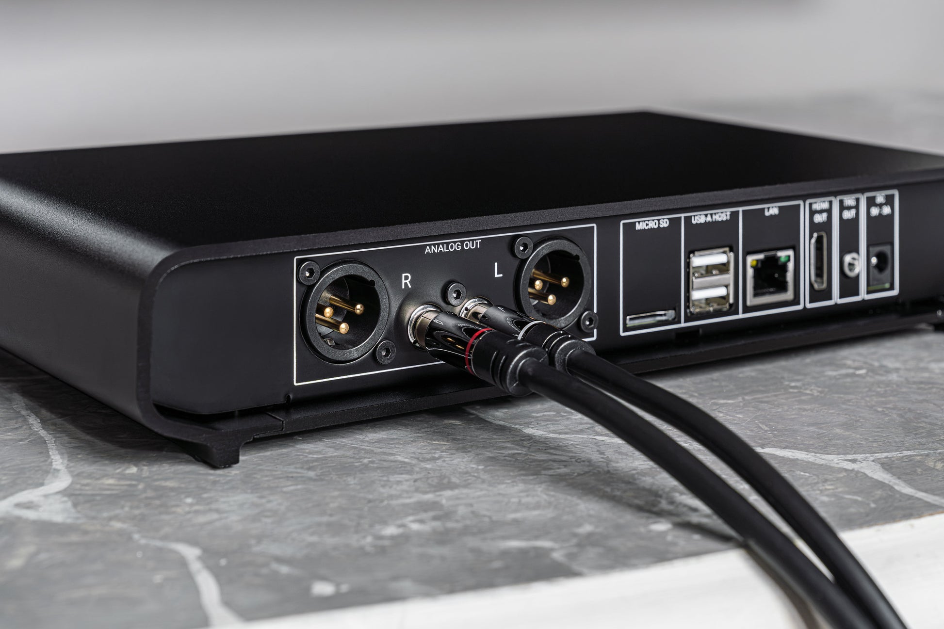 Close-up of the rear panel of a black audio device with XLR outputs, USB-A and USB-C ports, and connected audio cables, resting on a gray marble surface.