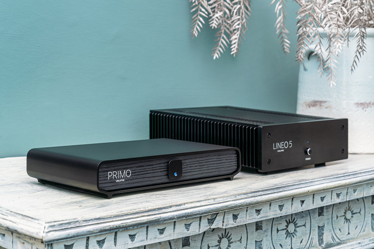 Two black audio equipment enclosures, labeled PRIMO and LINEO 5, displayed on a distressed white mantelpiece against a teal wall.