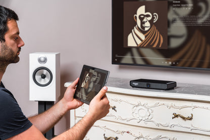 A man holds a tablet showing a projected image onto a wall-mounted screen above a white ornate cabinet, with a black projector on the cabinet and a white speaker beside him.