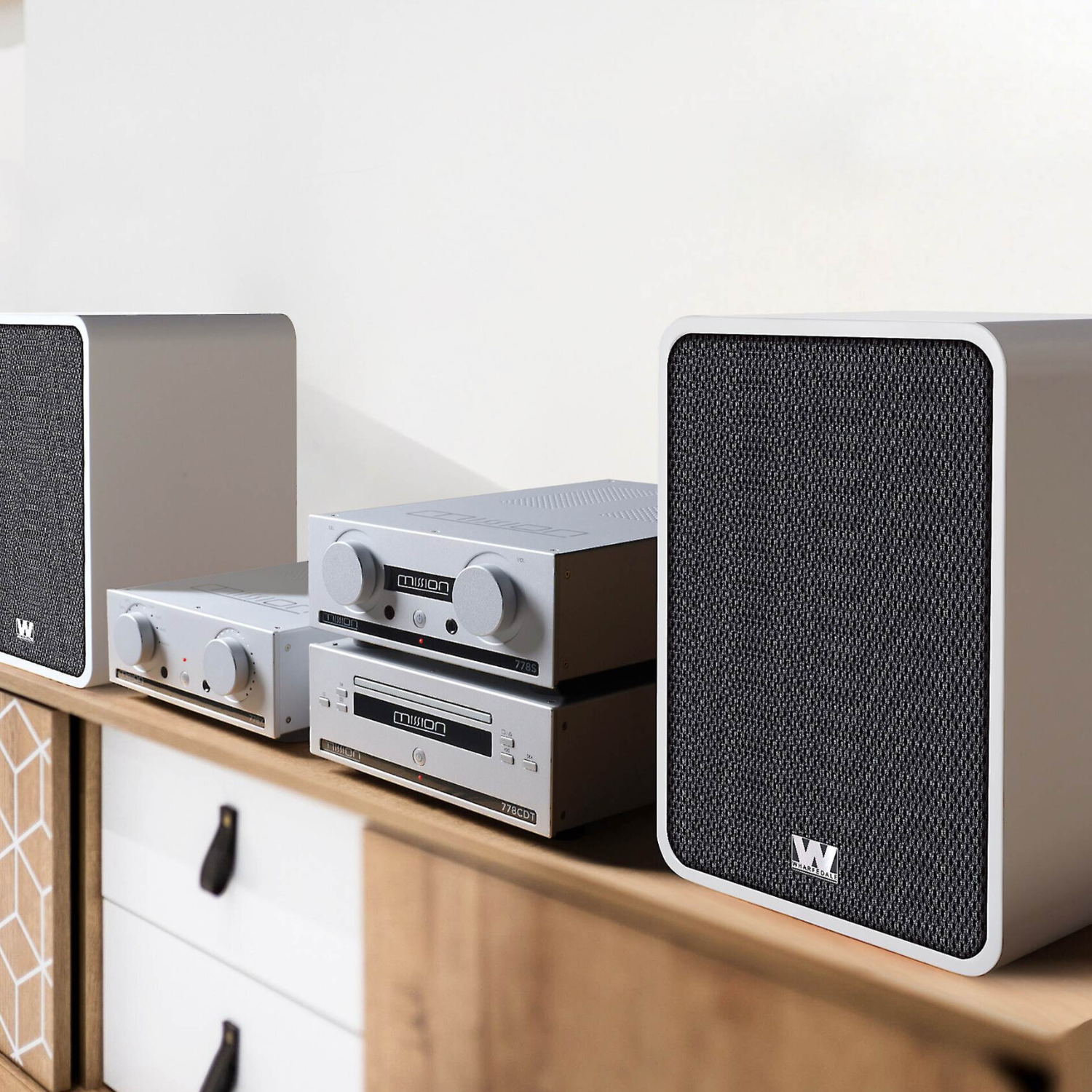 A pair of white Wharfedale Denton 1S speakers, and two silver receiver units stacked on a wooden cabinet against a white wall.