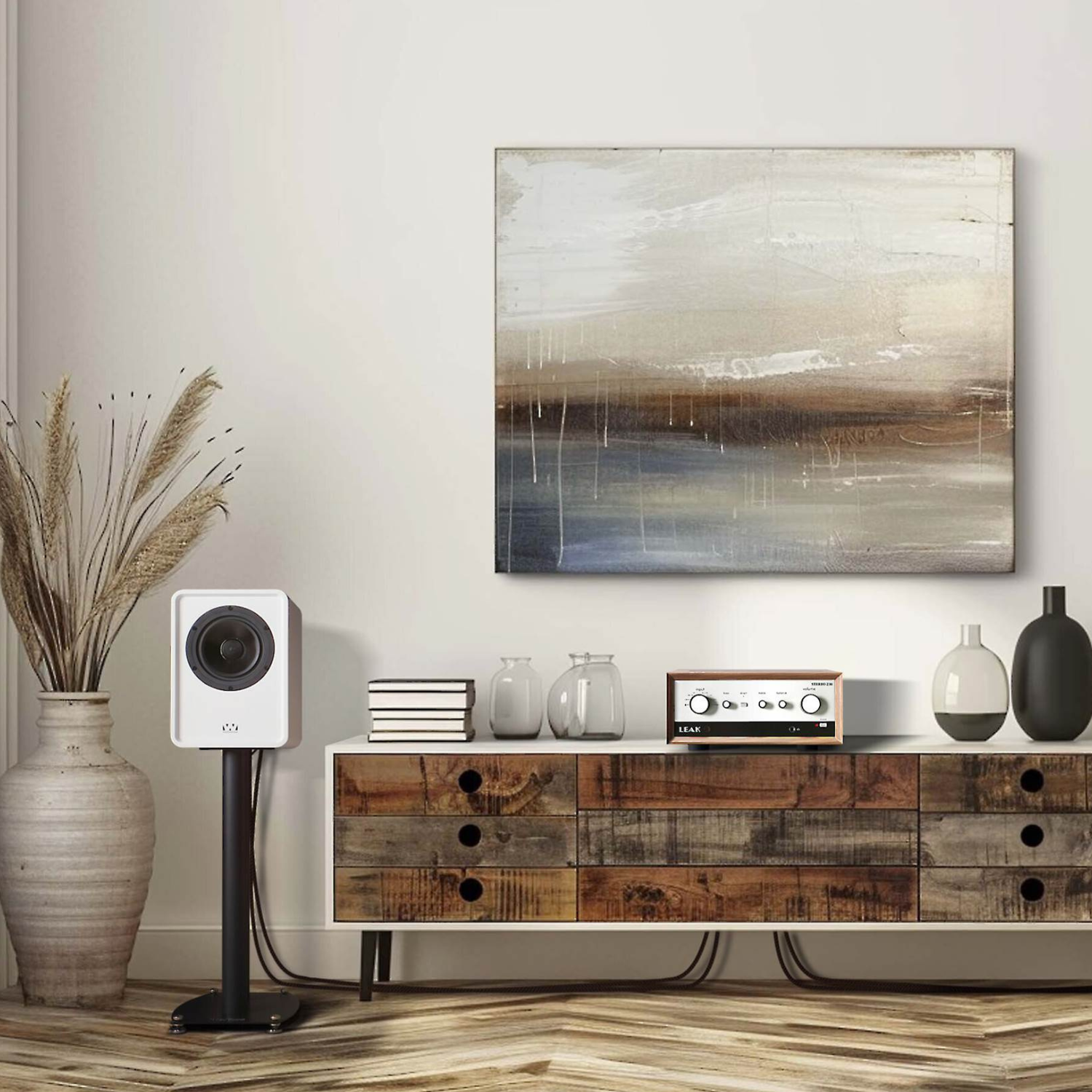 An abstract landscape painting with beige, brown, and blue tones hangs on a light wall above a rustic wooden console table styled with books, glass jars, a white speaker, and decorative vases.