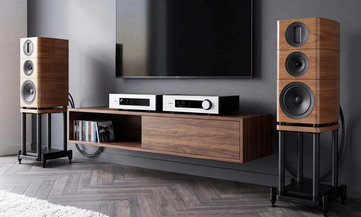 A pair of tall wood-encased speakers on black stands, flanking a floating wood TV console with a mounted wall screen and audio equipment in a modern room.
