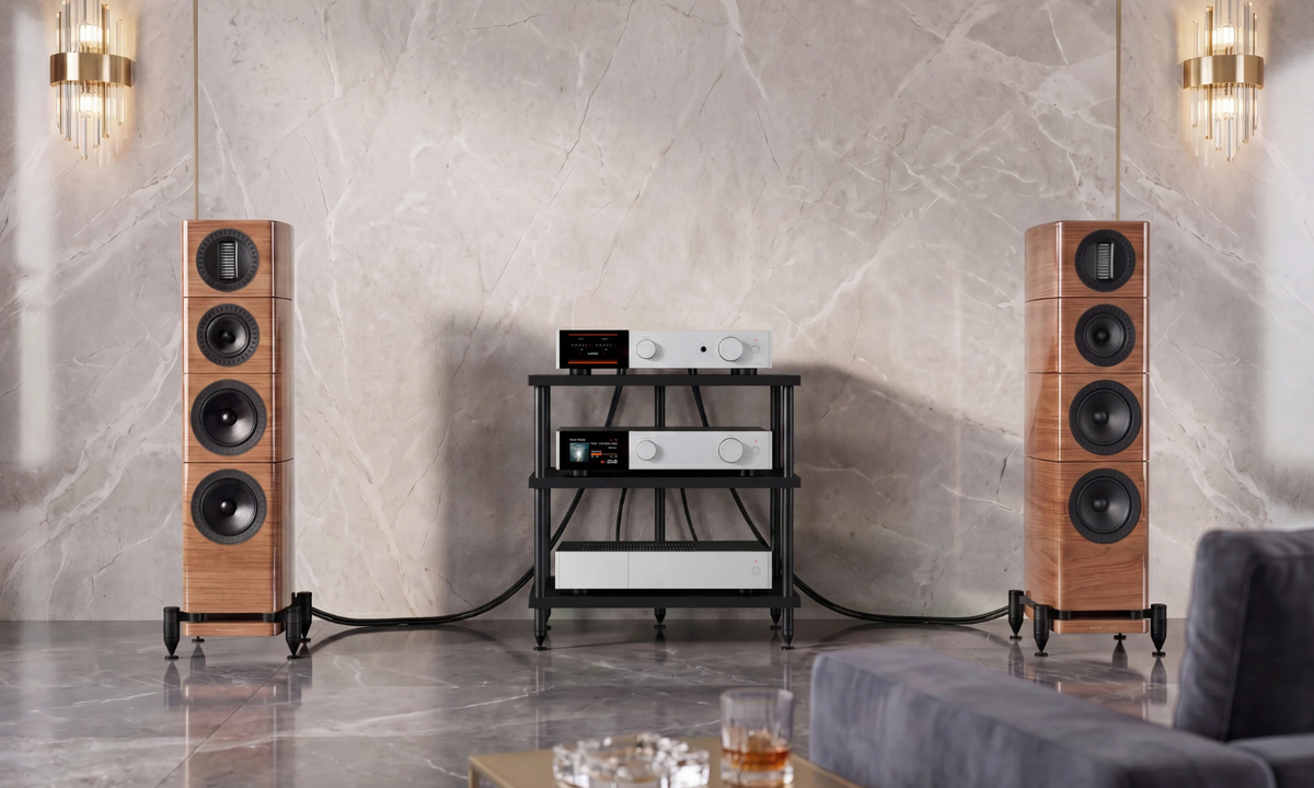 A high-end audio speaker system in a modern living room setting, featuring two tall wooden floor-standing speakers flanking a black and white media console against a marble wall.