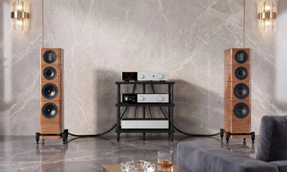 A high-end audio speaker system in a modern living room setting, featuring two tall wooden floor-standing speakers flanking a black and white media console against a marble wall.