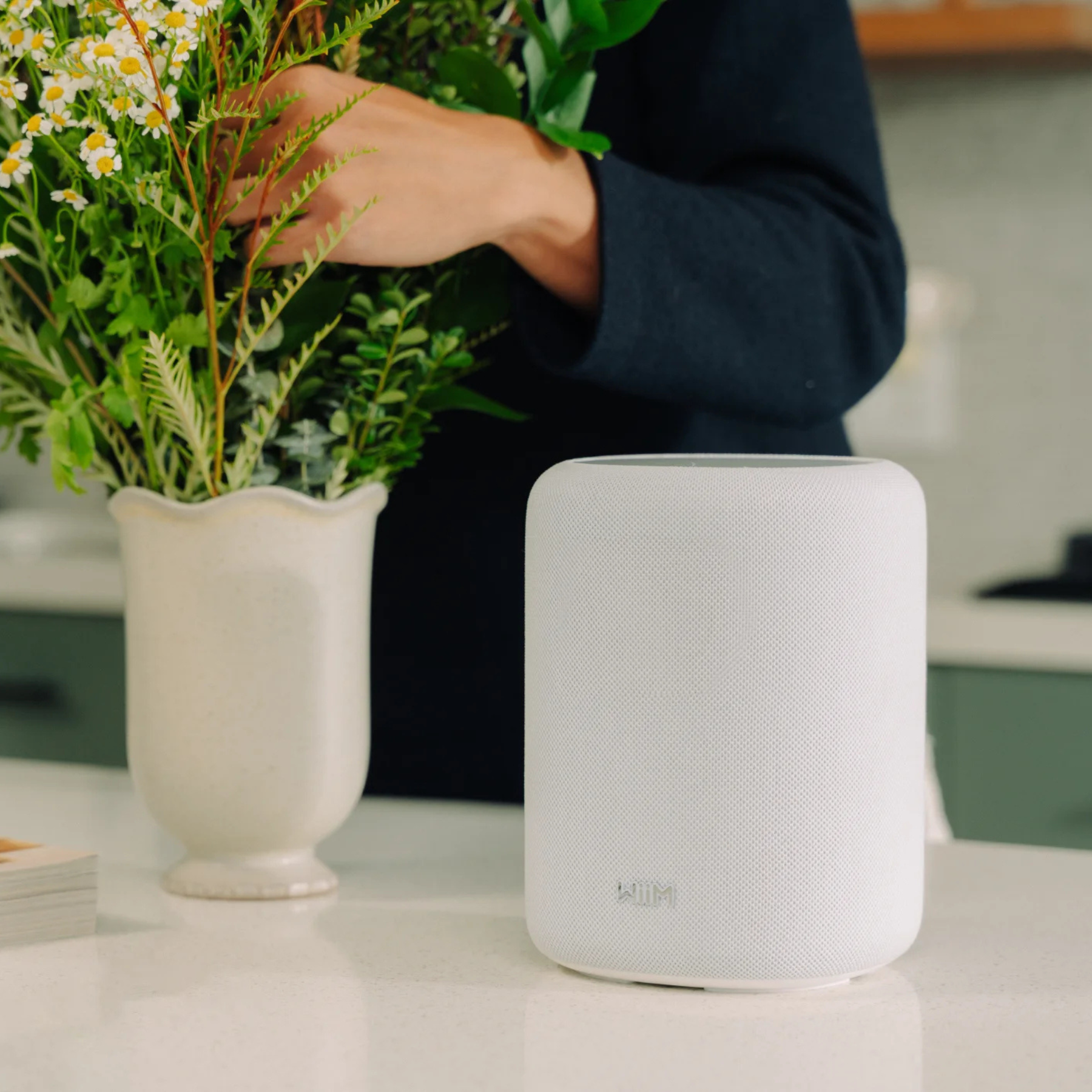 White smart speaker on a table with a vase of flowers and a person in the background