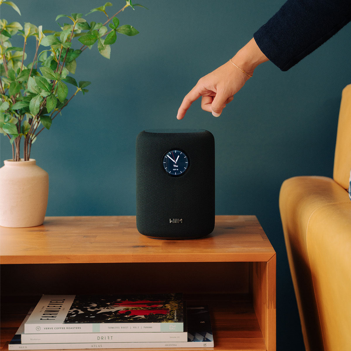 Person interacting with a black WiiM Sound Smart Speaker on a wooden shelf against a teal wall.