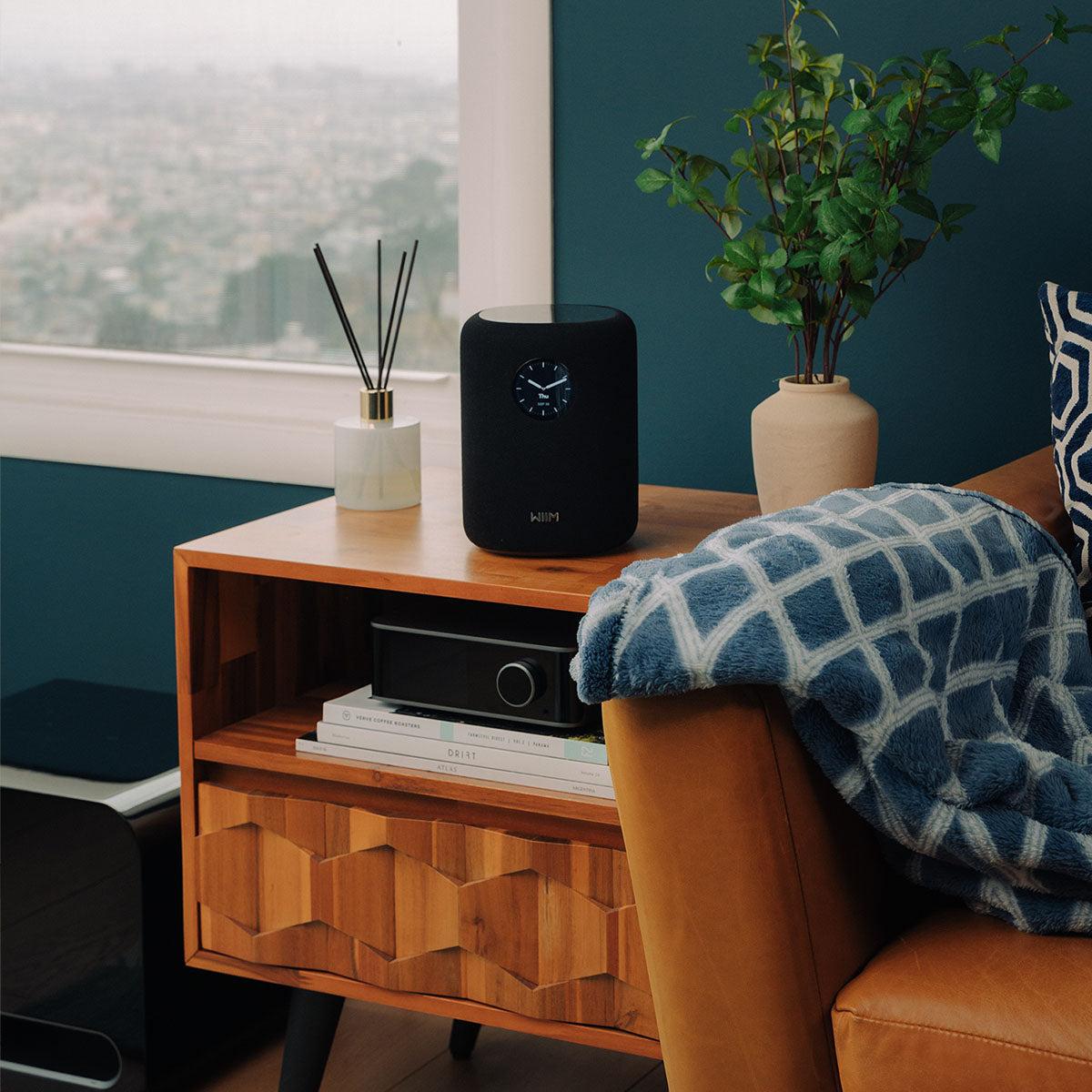 Wooden side table with a black WiiM Sound Smart Speaker, books, and a plant in a room.