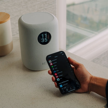 White WiiM Sound Smart Speaker on a surface with a hand holding a smartphone displaying a music app.