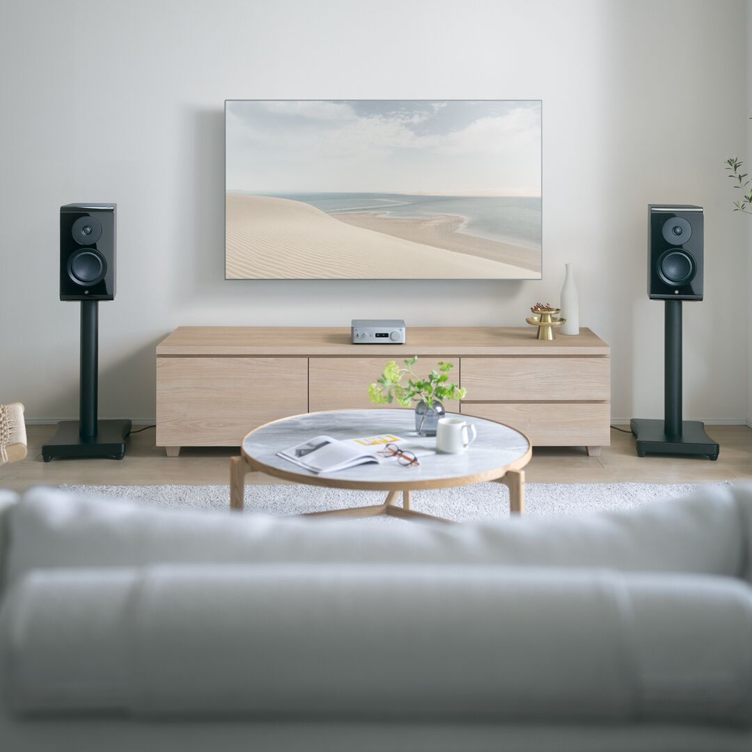 A modern living room setup featuring a large wall-mounted art print of a desert beach, flanked by two black speakers on stands above a light wood media console. A round marble-top coffee table sits in front, with a blurred white sofa in the foreground.