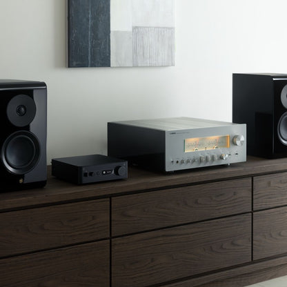 A silver and black audio receiver and amplifier system displayed on a dark wood media console, flanked by two black speakers against a light wall with abstract wall art.