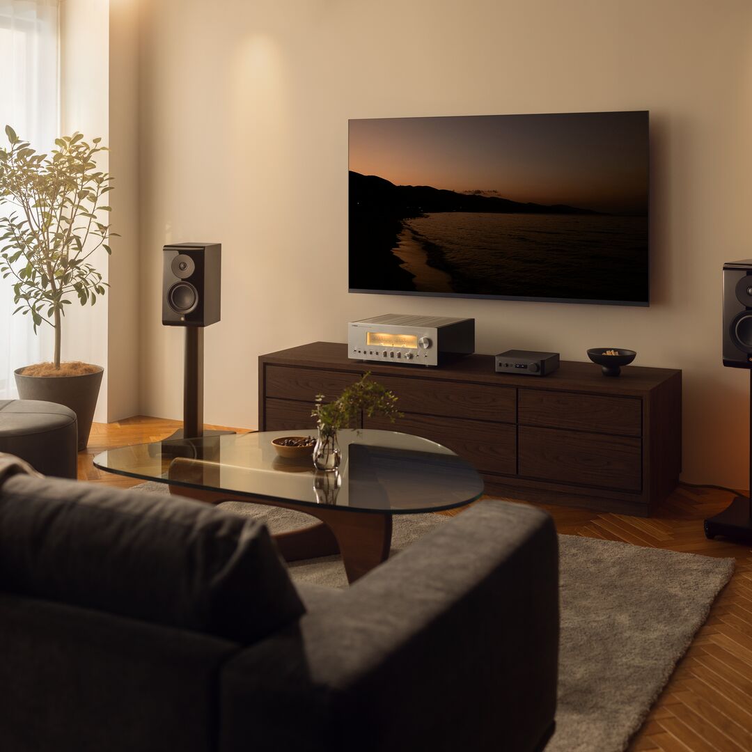 A modern living room setup featuring a large wall-mounted TV displaying a sunset seascape, paired with a dark wood media console, a silver audio receiver, and matching speakers on a wooden floor.