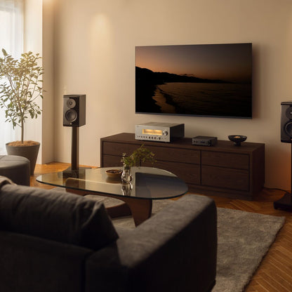 A modern living room setup featuring a large wall-mounted TV displaying a sunset seascape, paired with a dark wood media console, a silver audio receiver, and matching speakers on a wooden floor.