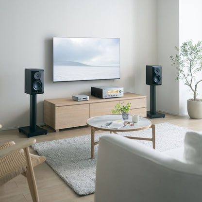 A modern living room setup featuring a pair of black bookshelf speakers on stands flanking a wall-mounted TV above a light wood media console, with a coffee table and rug in the foreground.