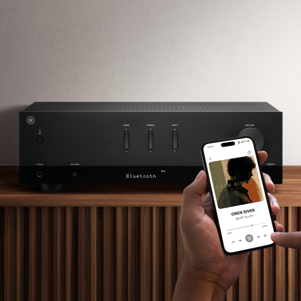 A black Bluetooth audio receiver sits on a wooden shelf against a textured light wall, with a hand holding a smartphone in the foreground showing a music player app.